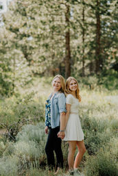 Two smiling women standing back-to-back holding hands in a sunlit pine-forest meadow, one in a white summer dress and the other in jeans and a floral top.