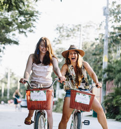 Two friends laughing on a summer bike ride with red wicker baskets along a sunlit, tree-lined tropical street, casual outfits and a straw hat