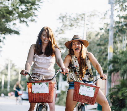 Two friends laughing on a summer bike ride with red wicker baskets along a sunlit, tree-lined tropical street, casual outfits and a straw hat