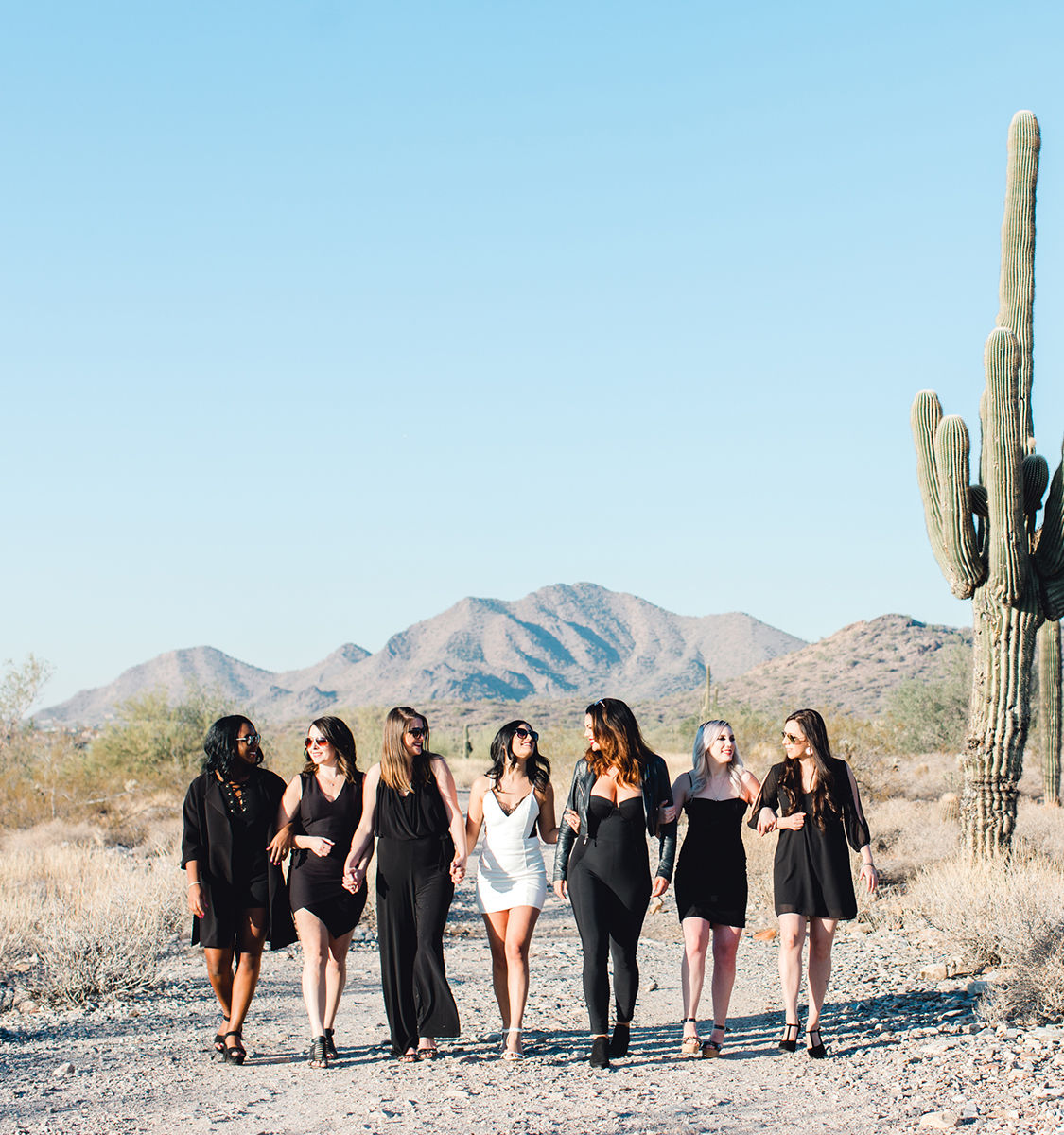 Seven women in black and white dresses walk arm-in-arm along a sunlit Arizona desert trail with a tall saguaro cactus and mountains under a clear blue sky.