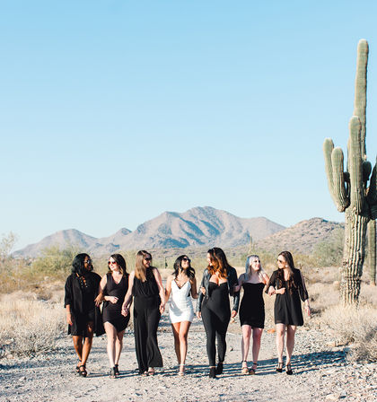 Seven women in black and white dresses walk arm-in-arm along a sunlit Arizona desert trail with a tall saguaro cactus and mountains under a clear blue sky.