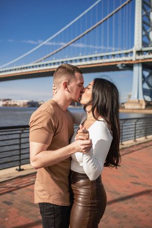 Couple kissing and holding hands on a sunny riverwalk with a large steel suspension bridge and blue sky in the background.
