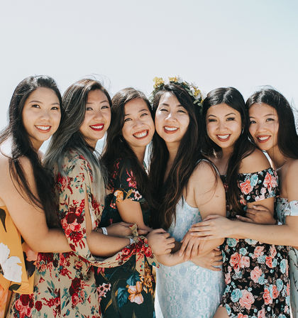 Six smiling women in colorful floral dresses hugging in a sunlit beach portrait, one wearing a flower crown — joyful group of friends outdoors.