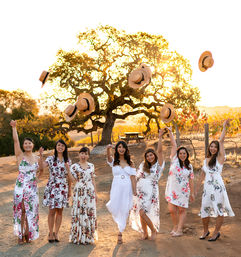 Seven women in white and floral dresses celebrate at a sunlit vineyard during golden hour, smiling and tossing straw hats into the air in front of a large oak tree.