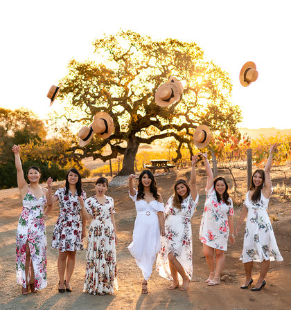 Seven women in white and floral dresses celebrate at a sunlit vineyard during golden hour, smiling and tossing straw hats into the air in front of a large oak tree.