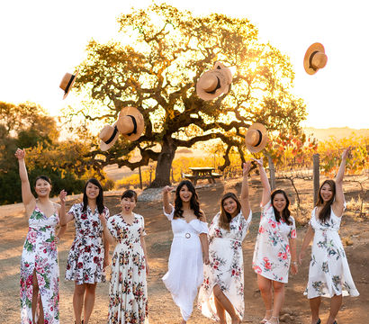 Seven women in white and floral dresses celebrate at a sunlit vineyard during golden hour, smiling and tossing straw hats into the air in front of a large oak tree.