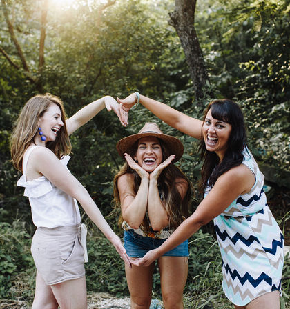 Three laughing friends in summer outfits in a sunlit woodland clearing forming a heart shape with their arms around a smiling woman wearing a straw hat