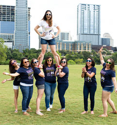Cheerful bachelorette party: eight women in matching tees and colorful sunglasses lift the bride-to-be in a white shirt above their shoulders in a sunny downtown park with a modern city skyline of skyscrapers behind them