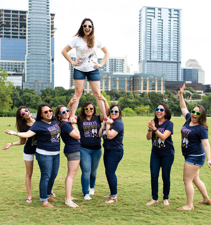 Cheerful bachelorette party: eight women in matching tees and colorful sunglasses lift the bride-to-be in a white shirt above their shoulders in a sunny downtown park with a modern city skyline of skyscrapers behind them