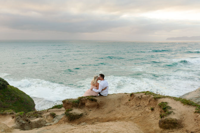 Romantic couple embracing on a rocky coastal cliff, overlooking crashing ocean waves and a soft pastel sky.