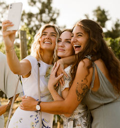 Three smiling women hugging and taking a selfie in a sunlit outdoor vineyard, carefree summer friends portrait