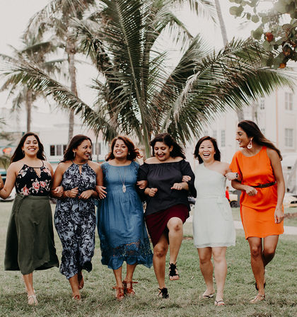 Six women friends linked arm-in-arm, laughing and walking across a grassy tropical park with tall palm trees and buildings in the background.