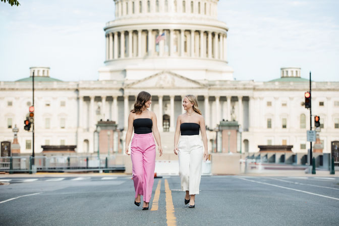 Two women in black tube tops and pink and white high-waist pants walking and smiling down a street toward the U.S. Capitol building in Washington, D.C.