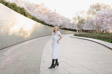Smiling woman in a white midi dress and black boots stands on a curved paved walkway lined with blooming cherry blossom trees and a low stone wall in a sunlit spring park.