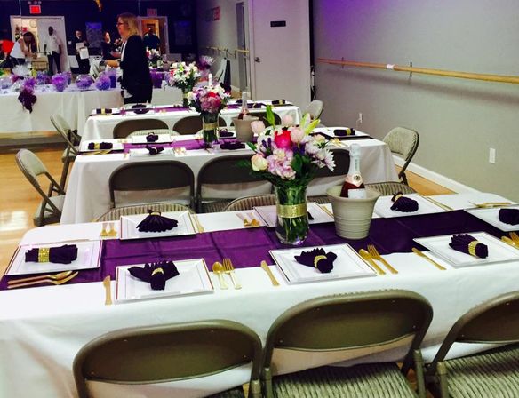 Long banquet tables in a dance studio set for a party with white tablecloths, purple runners and folded napkins, gold flatware, floral centerpieces and a chilled bottle in an ice bucket.