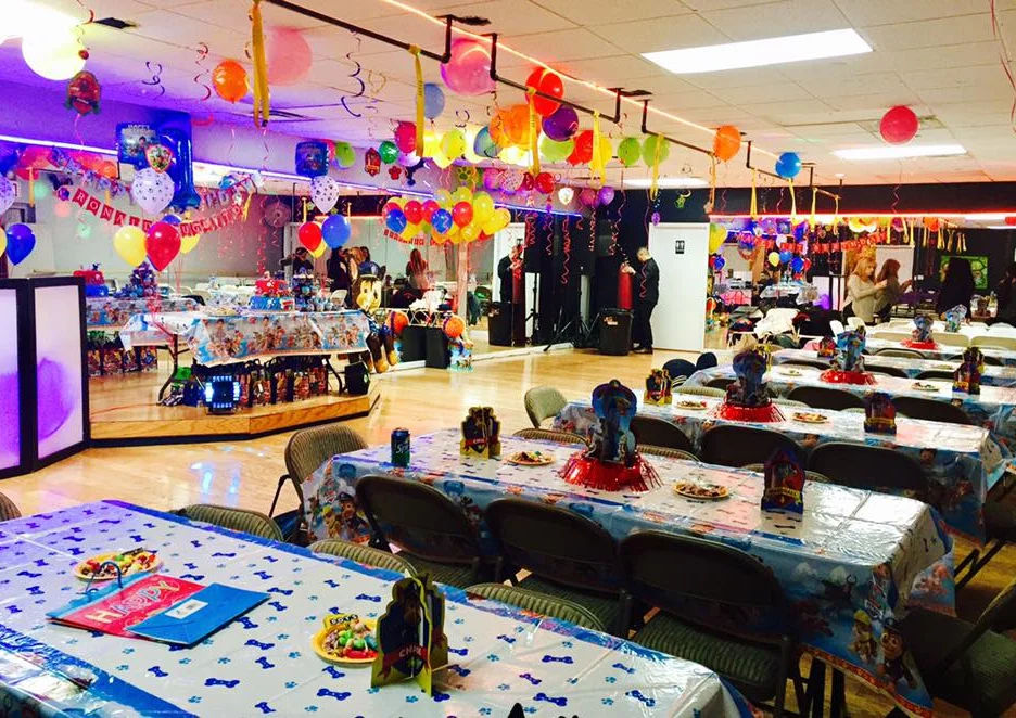 Bright indoor kids' birthday party room with long tables in themed tablecloths, colorful balloon bouquets, hanging streamers and a small dance floor.