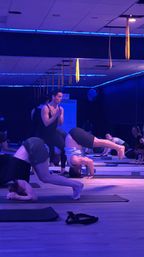 Glowing blue-lit indoor yoga studio class with participants on mats practicing headstands and core poses while an instructor guides them.