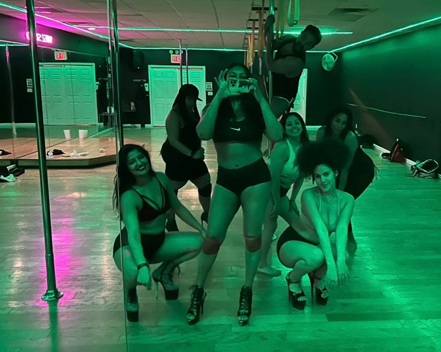 Mirror selfie of a pole fitness group posing in athletic wear on a wooden studio floor with chrome poles under green neon lighting.