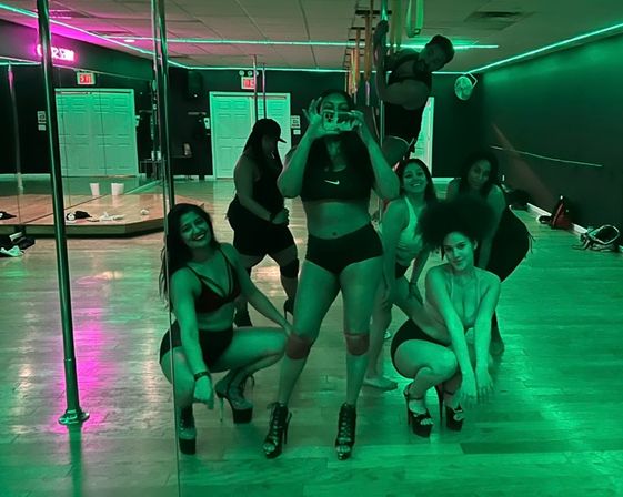 Mirror selfie of a pole fitness group posing in athletic wear on a wooden studio floor with chrome poles under green neon lighting.