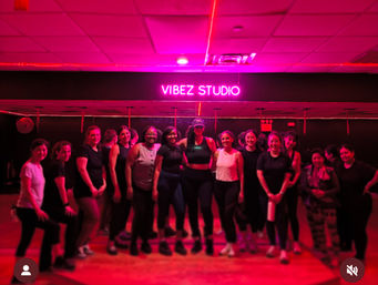 Group of women smiling after a high-energy group fitness class in a neon-pink lit indoor workout studio with a glowing sign overhead.