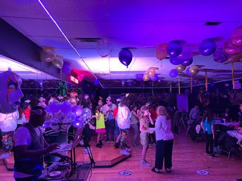 Purple-lit indoor celebration with balloons, DJ booth and a mixed-age crowd dancing on a small wooden dance floor