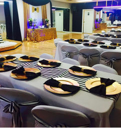 Indoor event hall set for a black, white and gold baby shower: long tables with striped runners, gold charger plates, black napkins and matching chair sashes in front of a gold-sequin dessert table.