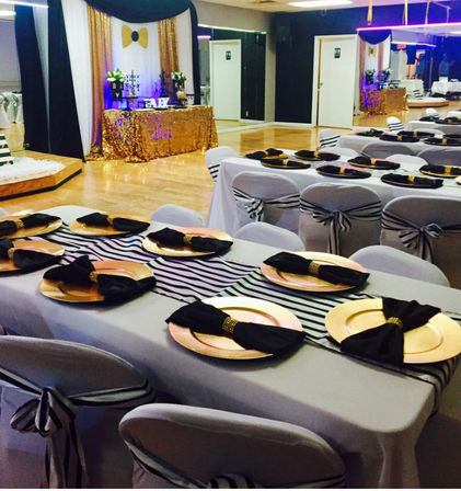 Indoor event hall set for a black, white and gold baby shower: long tables with striped runners, gold charger plates, black napkins and matching chair sashes in front of a gold-sequin dessert table.