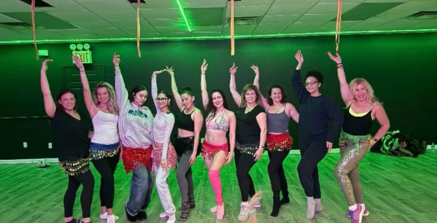 Group of women in colorful hip scarves striking raised-arm poses during a belly-dance class in a dance studio with green ambient lighting.