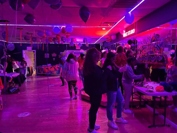 Neon-pink indoor celebration with balloons and streamers above a wooden dance floor, young guests mingling by tables of party props.