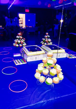 Neon blacklight party table in an indoor venue with a tiered cupcake stand, two rectangular sheet cakes, colorful glow bracelets and balloons on a blue tablecloth.