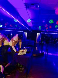 DJ at an indoor neon glow party adjusting a laptop and mixer under purple blacklight, fluorescent balloons hanging from the ceiling above a mirrored wall and glossy dance floor.
