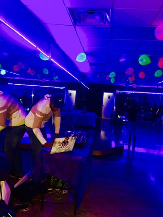 DJ at an indoor neon glow party adjusting a laptop and mixer under purple blacklight, fluorescent balloons hanging from the ceiling above a mirrored wall and glossy dance floor.