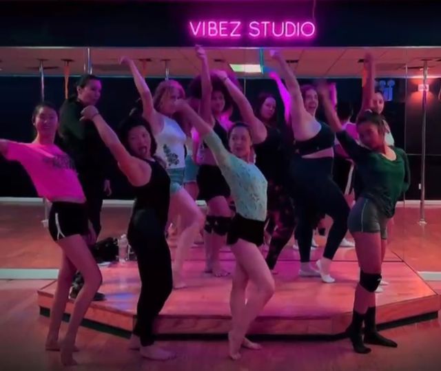 Energetic group of dancers striking poses on a small mirrored stage with poles under neon pink lighting in a dance studio, lively pole-dance class vibe.