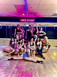 Vibrant neon-lit dance studio group: adults in athletic wear posing on and around a small stage and mirrored wall after a pole-fitness class, with wooden floors and one performer doing a full split in front.