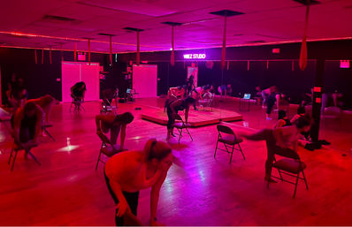 Neon-lit chair workout in a mirrored dance studio: participants stretch and balance on folding chairs under pink-red lighting on a hardwood floor.