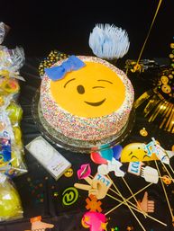 Emoji birthday cake with winking face and blue bow, covered in sprinkles on a decorated party table with emoji photo props, confetti, and colorful treats