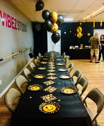 Emoji-themed black-and-gold party in a dance studio: long banquet table with black tablecloth, emoji plates and napkins, black and gold balloon centerpiece, folding chairs, wooden floor and wall-mounted barre.