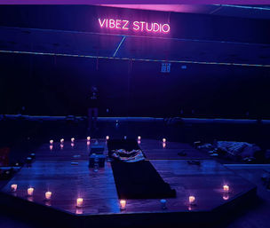 Candlelit indoor yoga/meditation studio with purple-blue neon glow, small candles arranged around a raised wooden platform, yoga mat and blankets ready for a calming session.