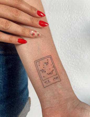 Tiny Polaroid-style line tattoo on inner forearm showing birds and stars labeled T.S. 1989, hand with red strawberry nail art