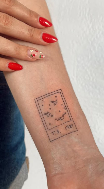 Tiny Polaroid-style line tattoo on inner forearm showing birds and stars labeled T.S. 1989, hand with red strawberry nail art