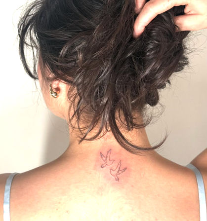 Close-up of a person's nape showing two tiny outline dove tattoos, dark hair in a messy bun and thin tank straps