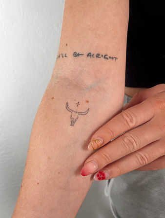 Close-up of inner forearm with a tiny minimalist bull skull tattoo and small cross above it, faint wrist script reading you'll be alright, and red ombré floral nail art on manicured fingers.