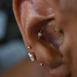 Close-up of a pierced ear featuring a gold star conch stud with a small clear gem and three tiny gold and clear gemstone studs along the lobe — trendy layered ear piercing and gold jewelry.