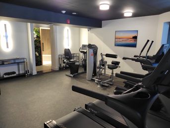 Bright modern gym with treadmills in the foreground, a recumbent bike and multi‑function strength machine in the center, padded benches and exercise stations along rubber flooring, wall-mounted sunset photo, soft overhead lighting and a doorway leading to a spa/sauna area.