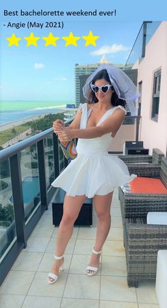 Bachelorette in a short white dress and veil popping champagne on an oceanfront hotel balcony with turquoise beach and high-rise skyline