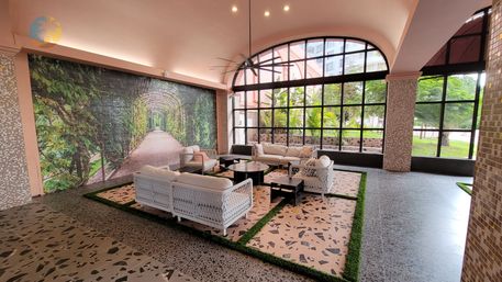 Sunlit tropical-inspired lobby lounge with white wicker sofas and black coffee tables on terrazzo floors, arched floor-to-ceiling windows framing palm trees and a large garden-arch mural
