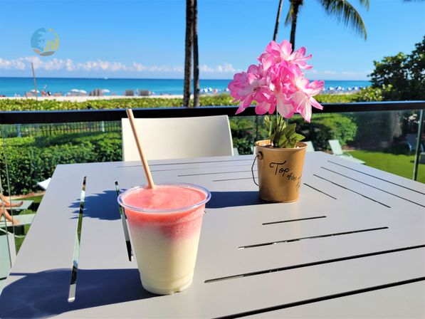 Sunny oceanfront balcony with a two-tone pink-and-white frozen drink on a gray table and a small potted pink flower, palm trees and sandy beach with blue ocean in the background.
