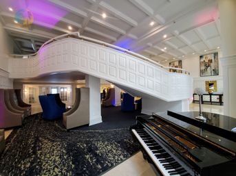 Elegant hotel lobby with sweeping white curved staircase, black grand piano in foreground, plush blue and gray lounge chairs, coffered ceiling and purple ambient lighting.