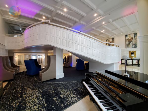 Elegant hotel lobby with sweeping white curved staircase, black grand piano in foreground, plush blue and gray lounge chairs, coffered ceiling and purple ambient lighting.