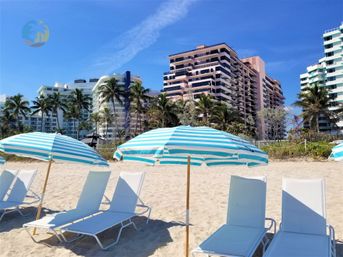 Sunny tropical beach with turquoise-and-white striped umbrellas and white lounge chairs on sand, palm trees and oceanfront high-rise condos under a bright blue sky